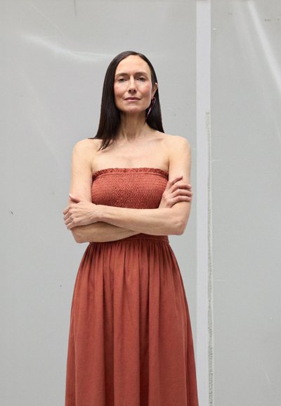 Mujer de cabello largo y oscuro con los brazos cruzados, vestida con un vestido sin tirantes de color óxido, frente a un fondo claro y liso.