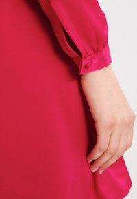 Fuchsia Kleid aus glattem, glänzendem Stoff, mit geknöpften Manschetten und einem lockeren Schnitt. Der Ärmel hat eine leichte Raffung am Handgelenk.