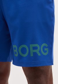 Μπλε αθλητικό σορτς φορεμένο στο πόδι ενός ατόμου με μεγάλα πράσινα γράμματα που σχηματίζουν το "BORG" κοντά στο τελείωμα στην αριστερή πλευρά.