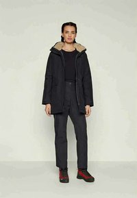 Moteris stovi vilkėdama juodą kapučiną paltą su smėlio spalvos pamušalu, juodas kelnes, juodą marškinius ir raudonus žygių batus prieš paprastą šviesų foną.