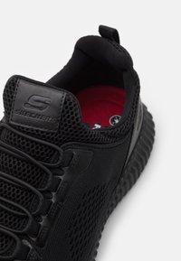 Black athletic shoe featuring a mesh upper, thick textured sole, elastic laces, and a red insole with Skechers logo on the heel.