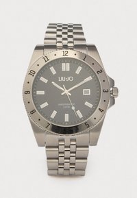 Ασημιές ανοξείδωτες ρολόγια με μαύρο καντράν, φωτεινές δείκτες ωρών και ένδειξη ημερομηνίας. Διαθέτει περιστροφικό στεφάνι και βραχιόλι συνδέσμων.