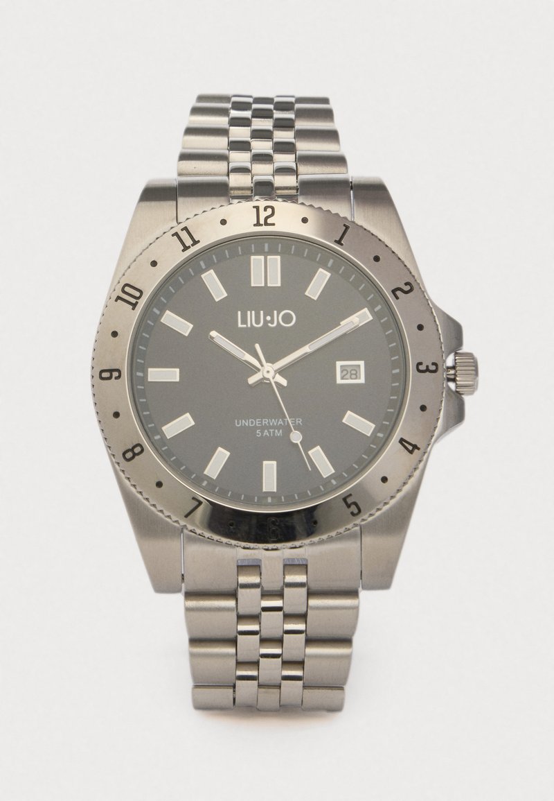 Ασημιές ανοξείδωτες ρολόγια με μαύρο καντράν, φωτεινές δείκτες ωρών και ένδειξη ημερομηνίας. Διαθέτει περιστροφικό στεφάνι και βραχιόλι συνδέσμων.