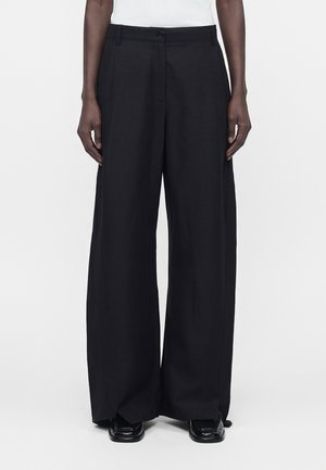 Modellen bærer sorte vidde bukser med knap og lynlås foran, kombineret med sorte sko, stående mod en ensartet hvid baggrund.