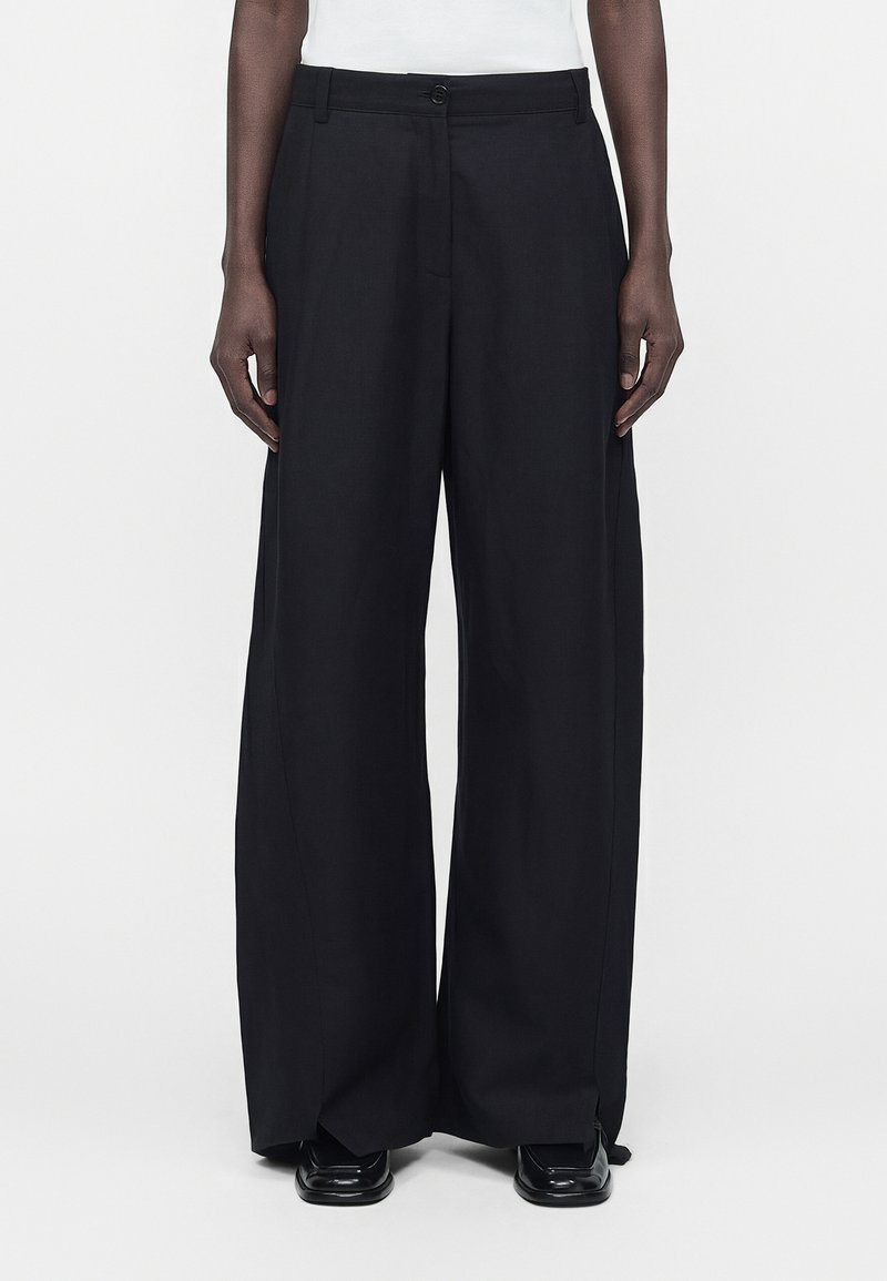 Το μοντέλο φοράει μαύρο παντελόνι με φαρδύ πόδι, κουμπί και φερμουάρ μπροστά, σε συνδυασμό με μαύρα παπούτσια, στέκεται μπροστά σε απλό λευκό φόντο.