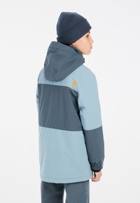 Veste à capuche bleu clair et bleu foncé avec des poignets élastiques et un logo sur l'épaule. Présente un design coloré en blocs et une coupe décontractée.