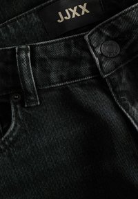 Κοντινό πλάνο μαύρου τζιν από denim που δείχνει θηλιά ζώνης, κουμπί με αρχικά της μάρκας και εσωτερική ετικέτα με το κείμενο JJJXX.