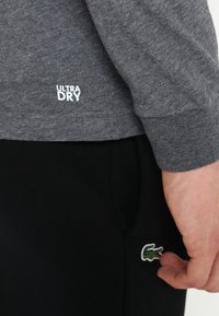 Graues langärmliges Hemd mit "ULTRA DRY" Aufschrift nahe dem Saum, kombiniert mit schwarzen Hosen, die an der Seite ein kleines grünes Krokodil-Logo tragen.