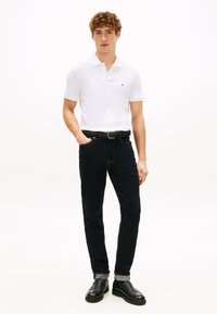 Fehér galléros póló kis logóval, sötétkék farmernadrág, fonott fekete öv és fekete, texturált felületű slip-on cipő.