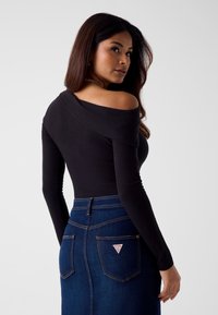 Zwarte off-shoulder top met lange mouwen, gecombineerd met een high-waisted donker denim rok met een driehoekig logo op het achterzakje.