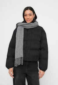 Μαύρο μπουφάν τύπου puffer με υφή, κομμένο σχέδιο, συνδυασμένο με απαλό γκρι κασκόλ με φουντωτά άκρα. Μοντέλο με σκούρο τζιν.