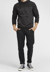 Sudadera negra con panel gris texturizado y capucha con cordón, combinada con pantalones negros. El modelo lleva zapatillas deportivas azul marino con suelas blancas.