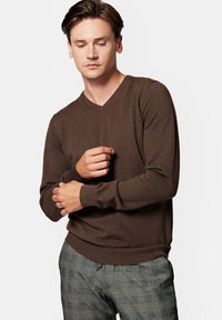 Brązowy sweter typu v-neck z dzianiny, z ściągaczami przy rękawach i dolnej krawędzi, noszony na tle spodni w kratę. Dyskretne logo na piersi.
