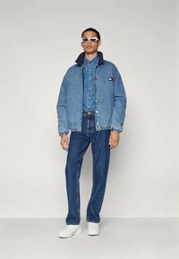 Casaco de denims com um tom azul, apresentando um colarinho azul-marinho em contraste e um logotipo. Combinado com jeans a condizer e ténis brancos. Óculos de sol angulares completam o visual.