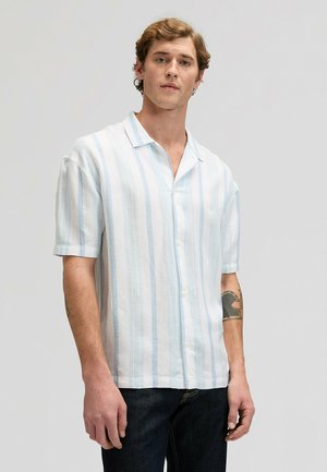 Junger Mann, der ein kurzärmeliges weißes Hemd mit hellblauen vertikalen Streifen und dunkle Jeans trägt, vor einem einfarbigen Hintergrund steht.