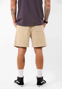 Shorts beige en tissu texturé, avec une poche arrière. Portés avec des baskets montantes noires et des chaussettes blanches.