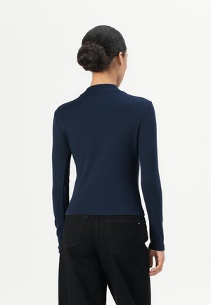 Femme avec les cheveux en chignon porte un haut bleu marine ajusté à manches longues et un pantalon noir, vue de dos sur un fond uni.
