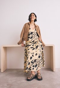 Robe maxi beige avec motifs tachetés noirs, superposée sous une veste en suède marron. Associée à des sandales marron épaisses. Accent avec un collier simple.