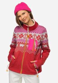Giacca in pile rossa con un design a fantasia caratterizzato da elementi bianchi e rosa, una tasca con zip rosa e zip e lacci di contrasto arancioni.