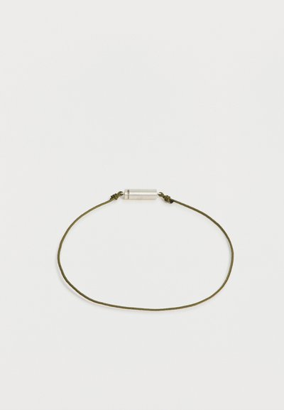 Bracelet fin en cordon vert olive avec un petit fermoir cylindrique en argent, posé à plat sur un fond blanc.