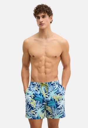 Uomo senza maglietta con capelli ricci che indossa pantaloncini da bagno blu con grandi motivi di foglie tropicali verdi, bianche e turchesi, mani in tasca.