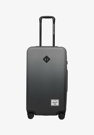 Valise rigide grise avec poignée télescopique, quatre roues et logo noir en forme de diamant au-dessus de l'étiquette blanche de la marque Herschel près de la base.