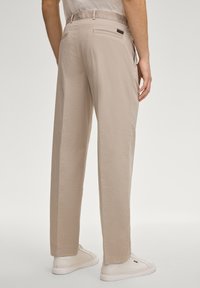 Pantalon beige clair en tissu lisse, coupe droite, poche arrière et étiquette de marque visible sur la ceinture.