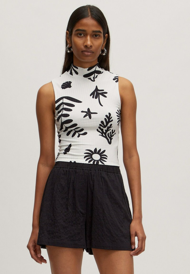 White sleeveless top with black botanical prints, high neckline, paired with black textured shorts; both made from lightweight materials.