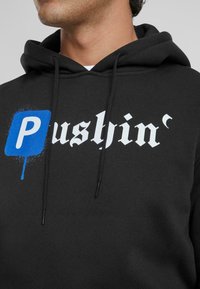 Felpa nera con cappuccio caratterizzata da un grande testo bianco "Pushin'" su uno sfondo effetto spray blu. Stringhe nere. Tessuto morbido.