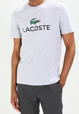 Mężczyzna w jasnoszarej koszulce Lacoste z zielonym logo krokodyla i czarnych kraciastych spodniach, stoi z jedną ręką w kieszeni.