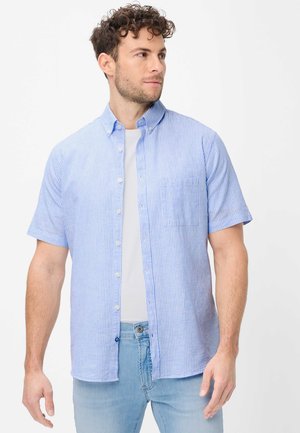 Junger Mann mit lockigem Haar, der ein blau-weiß gestreiftes kurzärmeliges Hemd über einem weißen T-Shirt und hellblauen Jeans trägt, schaut nach rechts.