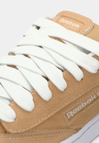 Sneaker scamosciato beige con lacci bianchi, caratterizzata da un logo Reebok sul lato e dettagli cuciti lungo il bordo.