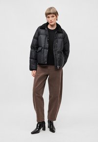 Črna napihnjena jakna z leskom in gumbnim zapenjanjem, z ovratnikom, stranskimi žepi, v kombinaciji z rjavimi hlačami in črnimi čevlji na peto.