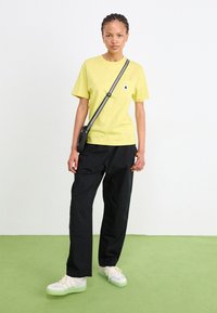 Yellow cotton t-shirt with a small pocket logo, paired with loose black pants and light shoes featuring a green sole. Simple, casual outfit.