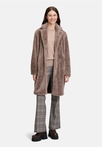 Kvinde iført en taupe falsk pelsfrakke, beige sweater, ternede bukser og chunky brune platformstøvler, smilende og stående mod en ensfarvet baggrund.