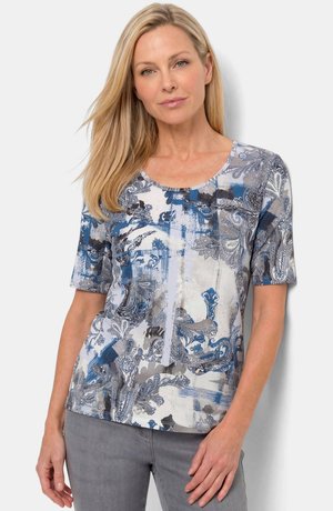 Blonde Frau trägt ein kurzärmeliges, blau-grau gemustertes Oberteil und graue Hosen, steht mit einer Hand entspannt an der Seite.