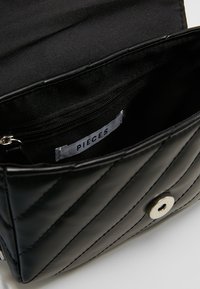 Borsa a tracolla nera trapuntata con patta aperta che mostra la tasca con zip interna e l'etichetta in tessuto con la scritta "PIECES."
