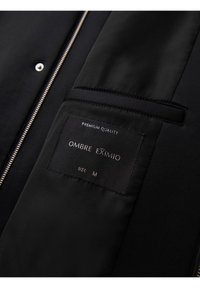 Gros plan sur la fermeture éclair d'une veste noire, la poche intérieure et l'étiquette en tissu indiquant "QUALITÉ PREMIUM OMBRE EXIMIO TAILLE M."