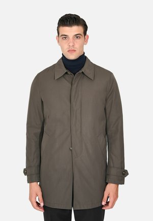 Uomo che indossa un cappotto verde oliva lungo fino al ginocchio con colletto e polsini con bottoni sopra un maglione nero a collo alto su uno sfondo semplice.