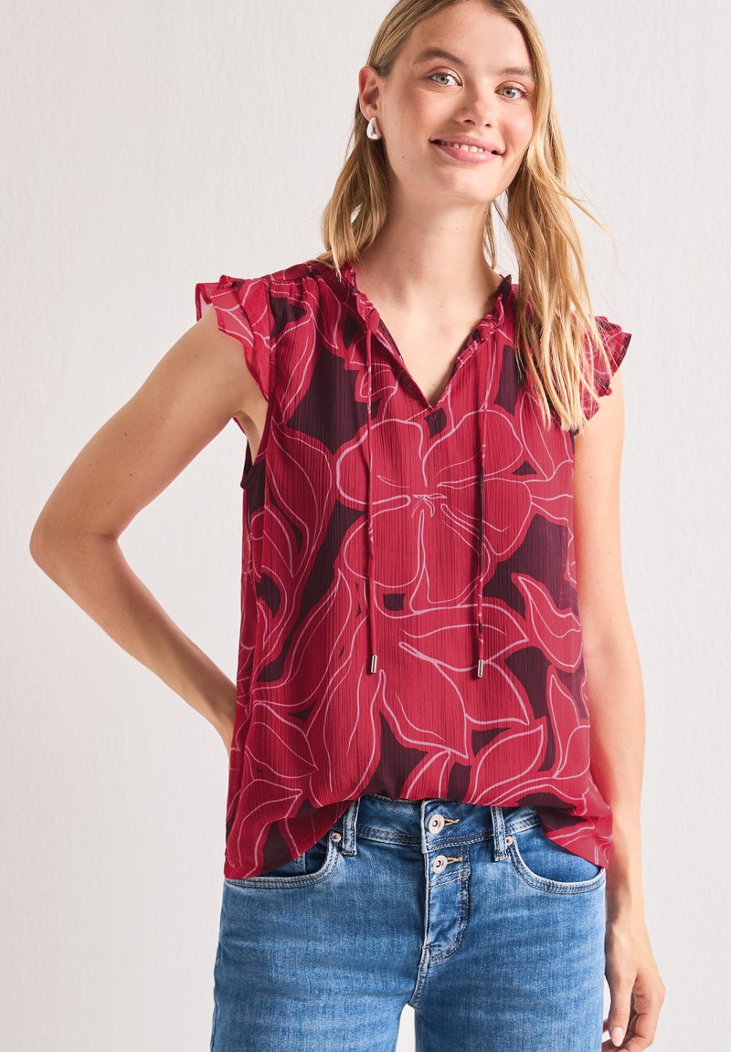 Jeune femme portant un chemisier rouge sans manches avec un grand motif floral et des manches à volants, associée à un jean bleu, souriant à la caméra.