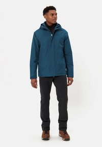 Eglė žalios spalvos neperšlampama striukė su gobtuvu, su užtrauktuku priekyje ir šoninėmis kišenėmis, derinama su juodomis kelniomis ir ruddais žygių batais.