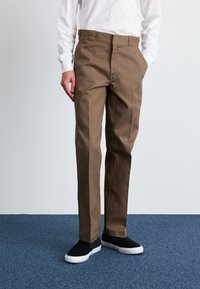 Brown straight-leg trousers made of fabric with a smooth texture. Features side pockets and is paired with black slip-on shoes on a blue carpet.