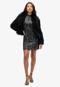 Veste en fausse fourrure noire sur une robe courte noire à sequins avec un motif ondulé. Présente un col haut et des talons noirs à lanières.