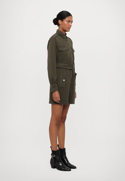 Rochie utilitară de culoare verde măsliniu, cu mâneci lungi, două buzunare la piept, talie împodobită cu curea și croi drept. Asortată cu cizme negre până la gleznă.