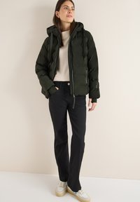 Giubbotto imbottito verde scuro con cappuccio, cerniere e corde regolabili, abbinato a un maglione leggero e pantaloni neri a zampa, indossando sneakers bianche.
