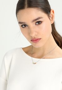 Silversmykke med cirkulære og geometriske vedhæng i guld- og sølvtoner. Modellen bærer en hvid top med en bred halsudskæring.