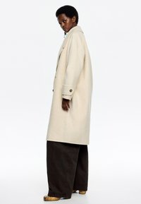 Cappotto in lana beige con vestibilità rilassata, grande colletto e polsini abbottonati, abbinato a pantaloni ampi scuri e scarpe con motivo.