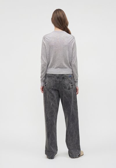 Femme aux longs cheveux bruns portant un pull gris clair et un jean noir délavé ample, debout de dos sur un fond blanc uni.