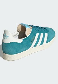 Tirkīza zamšādas apavi ar baltiem svītrojumiem, teksturēta gumijas zole un krēmkrāsas akcenti. Sānos ir zelta "GAZELLE" zīmols.