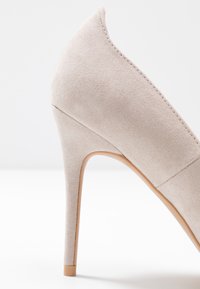 Zapato de tacón alto beige hecho de suave gamuza, con punta afilada y un delgado tacón tipo estilete. Textura suave con detalles de costura limpios.